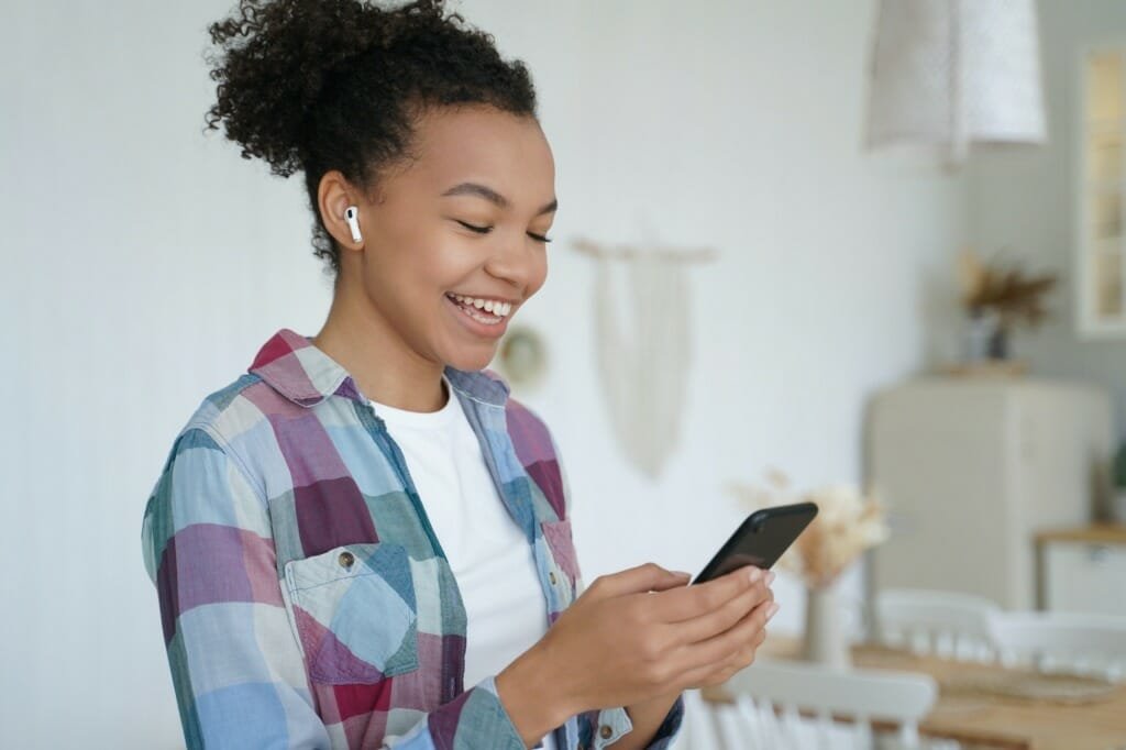 African american teen girl in earphone hold smartphone, listen audio message, use music apps at home