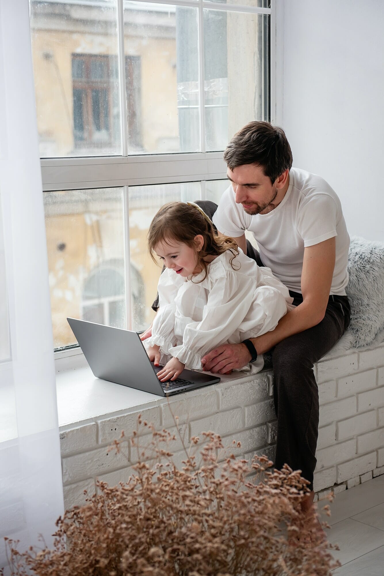 Father and daughter are playing on laptop, sitting on wooden, white windowsill near the window, Video Game Development Studio