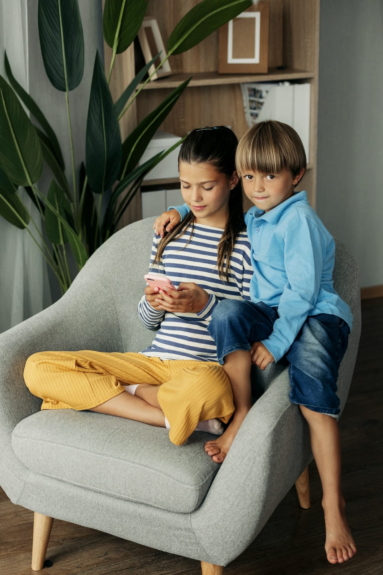 Sister and younger brother are sitting at home in an armchair and playing on the phone