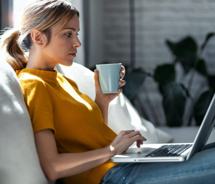 Confident woman working with her laptop while drinking a cup of coffee sitting on a couch at home