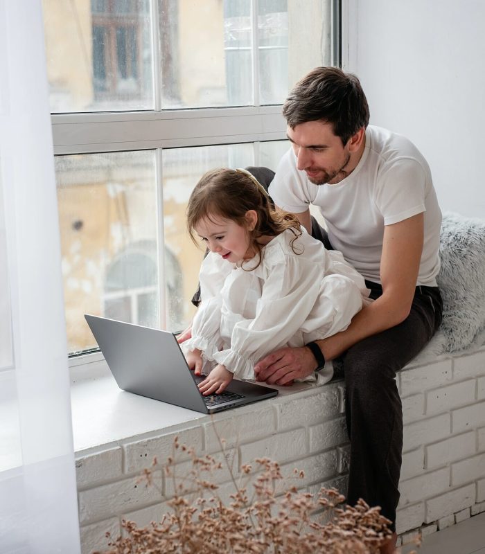 Father and daughter are playing on laptop, sitting on wooden, white windowsill near the window, Video Game Development Studio