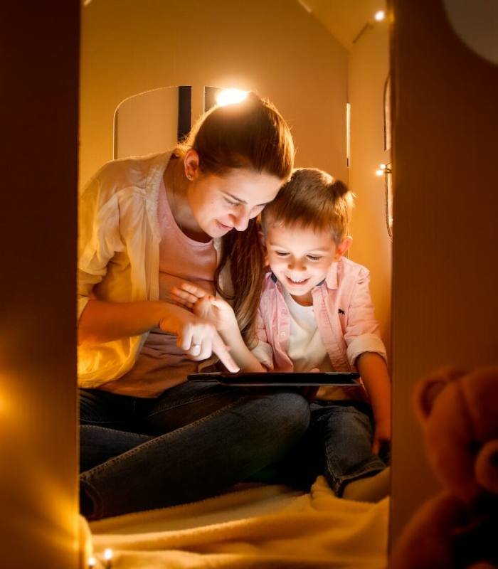 Happy laughing boy with young mother playing games and using tablet computer at night before going