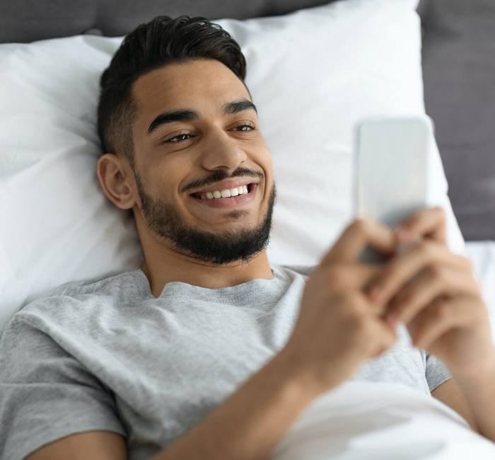 Mobile Gaming. Cheerful Young Arab Man Using Smartphone While Lying In Bed