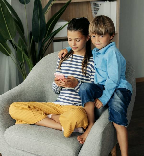 Sister and younger brother are sitting at home in an armchair and playing on the phone