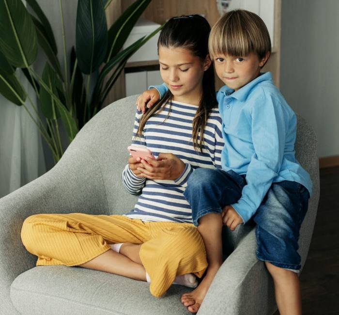 Sister and younger brother are sitting at home in an armchair and playing on the phone