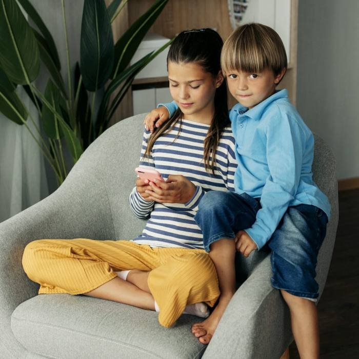 Sister and younger brother are sitting at home in an armchair and playing on the phone