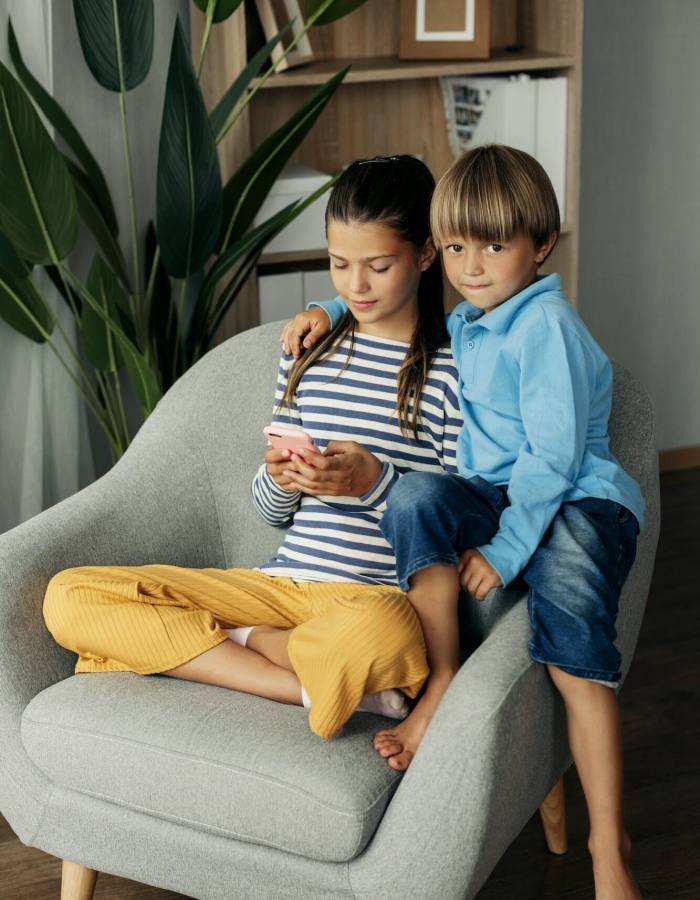 Sister and younger brother are sitting at home in an armchair and playing on the phone